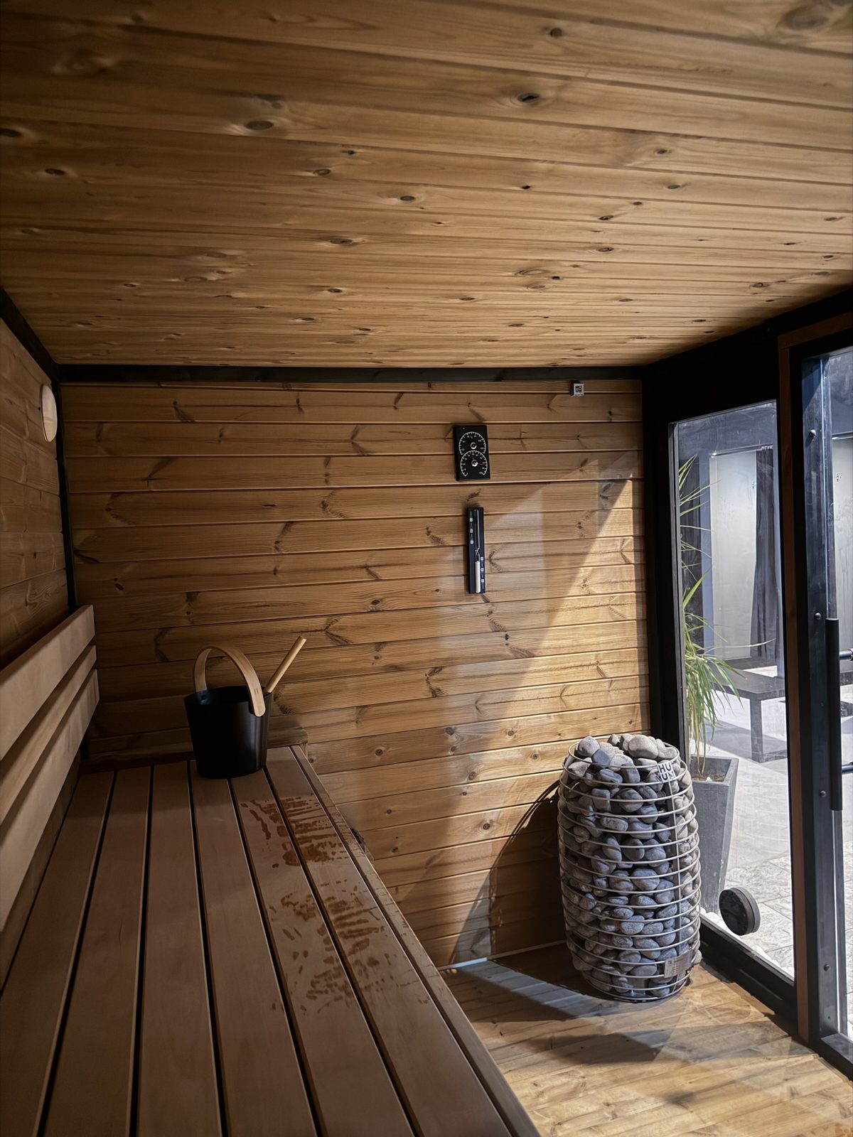 Inside the Finnish sauna at RE:UP Altrincham: wooden bench, water bucket and ladle, and the stone heater beside the glass door