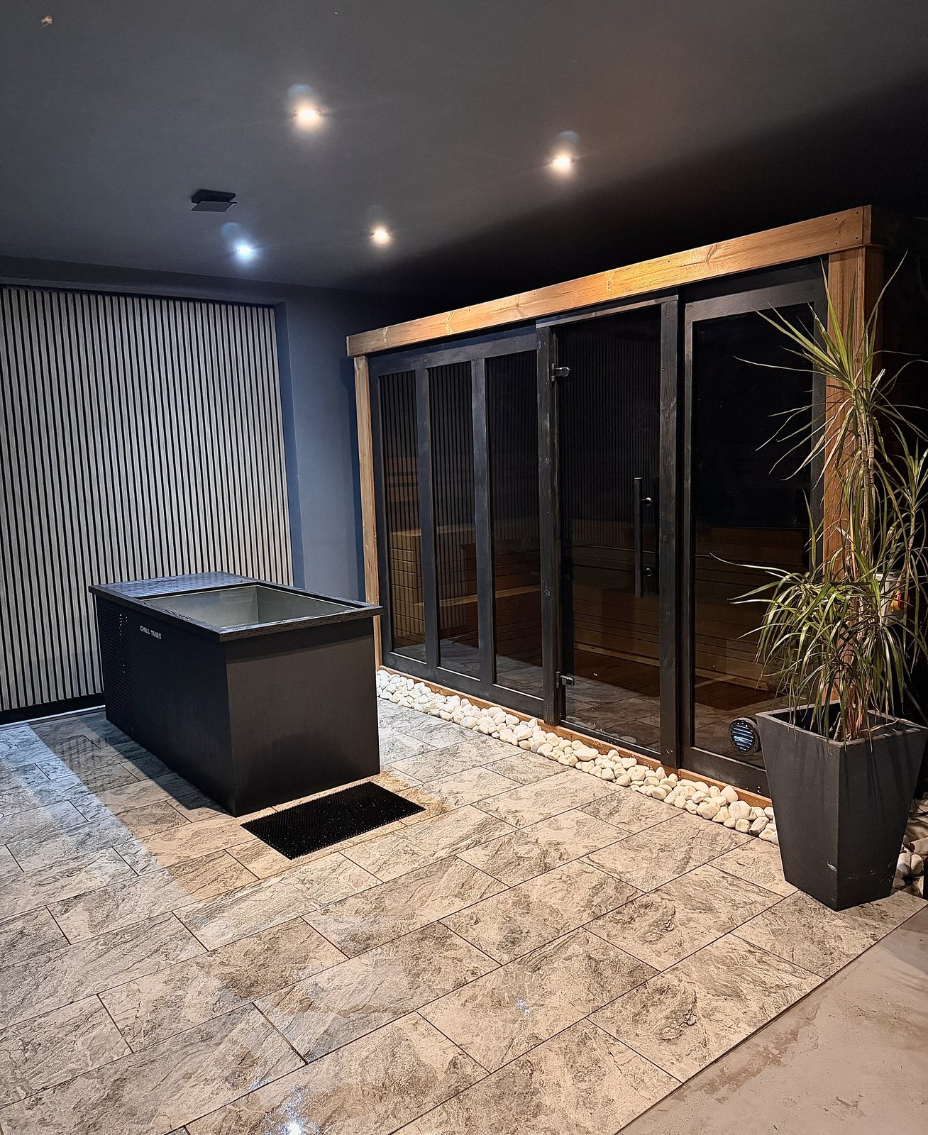 A cold plunge ice bath in front of the Finnish sauna cabin at RE:UP Altrincham, with the glass doors and a plant beside it