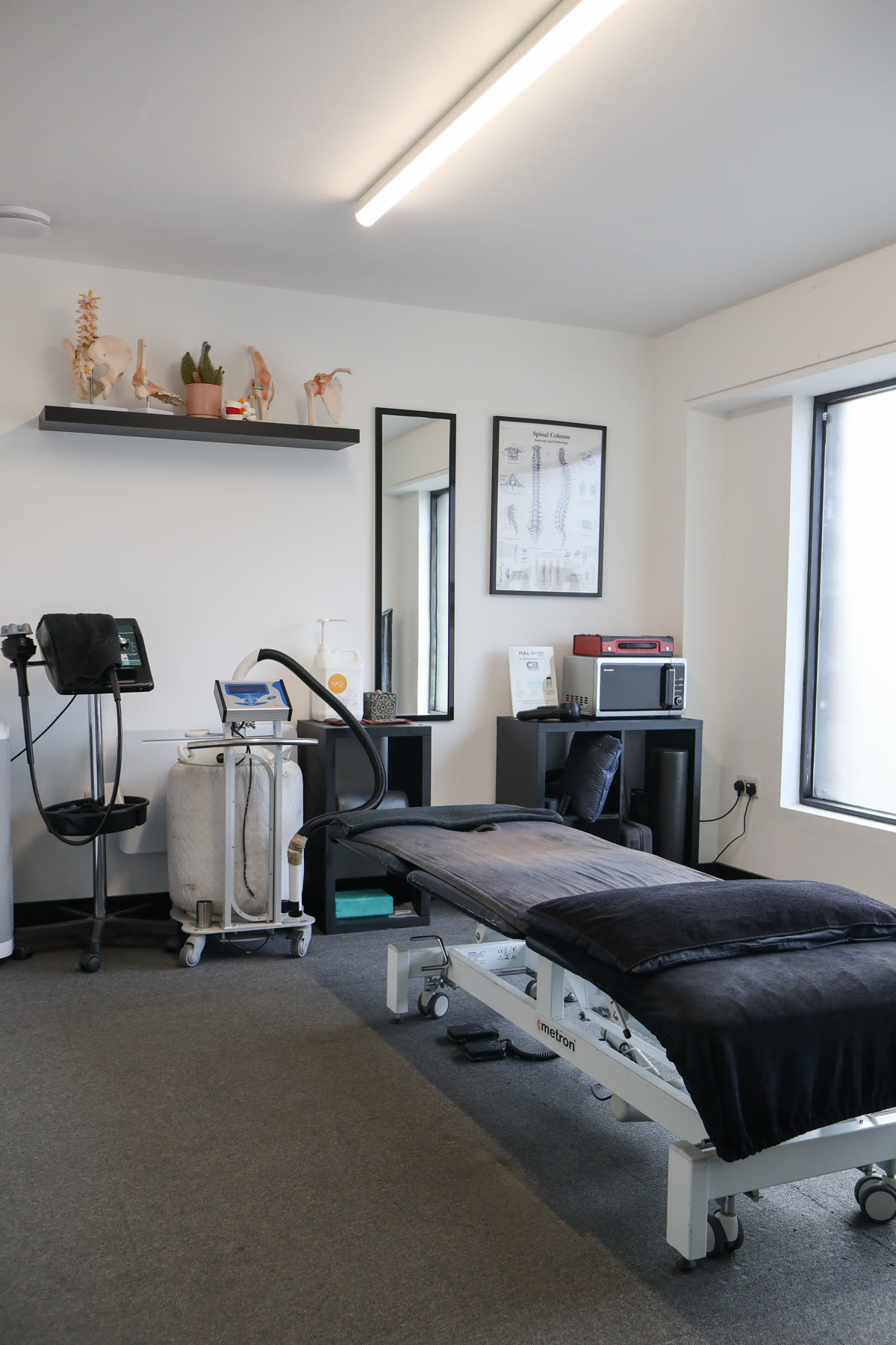 A treatment room at RE:UP Altrincham: an electric treatment couch with a dark cover, a shockwave therapy unit on a trolley, and a shelf of anatomy models under the window